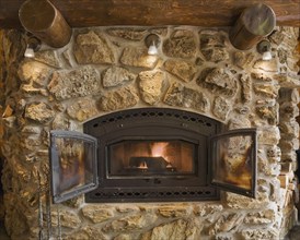 Lit fieldstone and porous rock fireplace in living room inside handcrafted red cedar log cabin