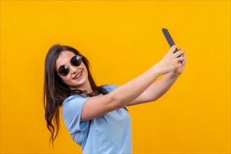 Studio portrait with yellow background of a chic woman with sunglasses taking a selfie smiling at