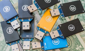 A collection of USB hardware wallets with Bitcoin logos spread over US dollar bills, in South Korea