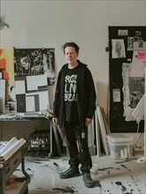 Individual in a graphic tee stands in a cluttered, artistic workspace with expressive intensity, AI