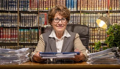 A smiling older woman in an office, surrounded by papers and a full bookshelf, symbolising
