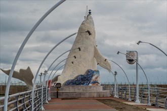 A unique sculpture stands at the end of a pier lined with arching street lamps on a cloudy day, in
