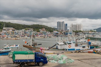 Fishing boats docked in a harbor with a truck and nets in the foreground, under an overcast sky, in