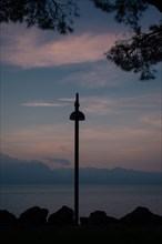 Silhouettes of a street lamp and branches against a twilight sky, mountains and Lake Garda in the