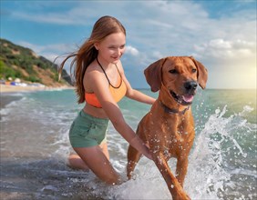 A girl plays with a dog, Rhodesian Ridgeback, on the beach and runs through the splashing sea