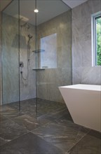 Clear glass shower stall and white freestanding vessel shaped bathtub in bathroom with grey ceramic