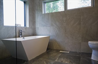 White freestanding vessel shaped bathtub in bathroom with grey ceramic tile floor and walls on