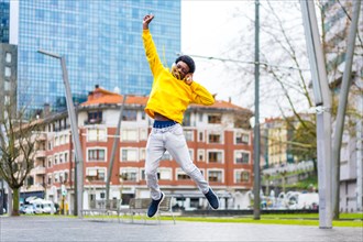 Happy young african man jumping while listening to music using headphones in the city