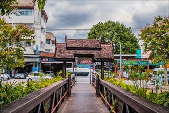 Traditional wooden bridge over a canal, with lush foliage and a city backdrop, in Chiang Mai,