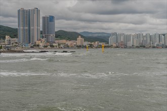 Overcast view of a city coastline with choppy sea, skyscrapers, and distant hills, in Ulsan, South