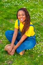 Vertical portrait of a cute african young woman in dungarees sitting on a park smiling at camera