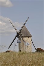 A stone windmill under a blue sky with clouds, surrounded by grass and multiple flags, Moulin du