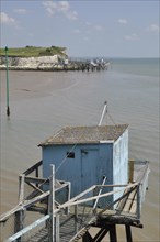 A tranquil beach scene with a blue hut on a wooden pier, under a sunny sky with a calm sea and