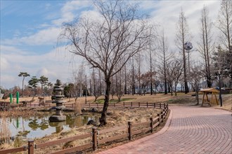A peaceful park scene with a pond, leafless trees and a brick footpath under a cloudy sky, in South