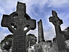 Two Celtic crosses in front of ancient ruins under a cloudy sky, possibly in Ireland