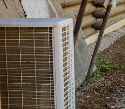 Closeup of air conditioner condenser unit on ground with exterior wall of log cabin in background