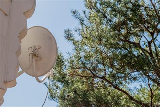 A white satellite dish mounted on a wall with a tree and blue sky in the background, in South Korea