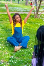 Vertical photo of an African young woman wearing dungarees sitting with arms raised while friend