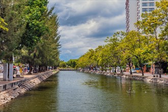 A serene urban canal lined with trees and a walking path under a cloudy sky, in Chiang Mai,