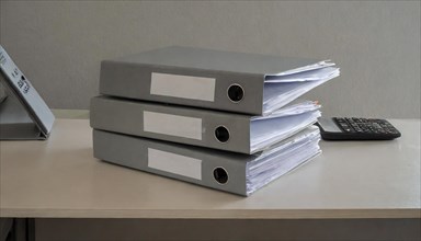Stacks of documents in grey file folders on a desk next to a calculator, symbolism bureaucracy, AI