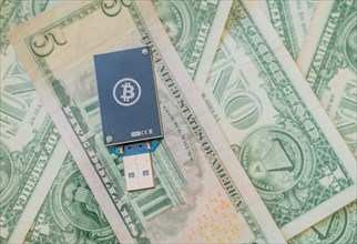 A dark blue USB hardware wallet with a Bitcoin logo on a background of US dollar bills, in South