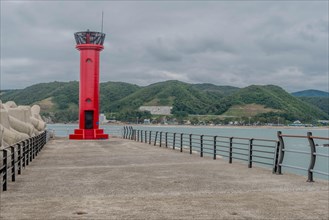 A vibrant red lighthouse stands on the pier with ocean and hills in the background, in Ulsan, South