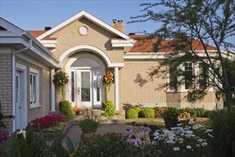 Tan brick home with white trim and landscaped front yard with raised border with red Pelargonium,