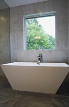 White freestanding vessel shaped bathtub in bathroom with grey ceramic tile floor and walls on