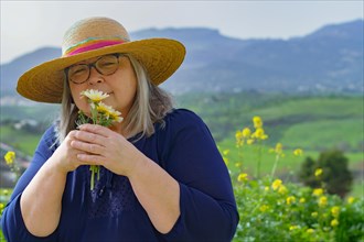 Mature woman with white hair and hat holding a bouquet of daisies in her hands in the background a
