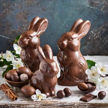 Three chocolate bunnies next to chocolate eggs and cinnamon sticks, surrounded by fresh flowers,
