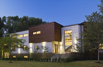 Illuminated beige stone with brown cedar wood siding modern cubist style home with landscaped front