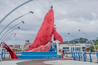Red sculpture dominating the harbor space with boats and blue railings under a cloudy sky, in