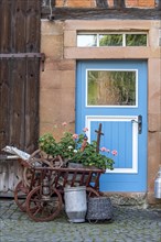 Curious decoration on a front door, flower pot, geraniums, old wagon with firewood, milk can and