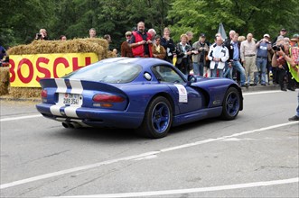 A blue sports car with a racing number and white stripes drives along a race track with spectators,