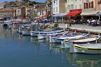 Cassis, the harbour, people strolling along a promenade with boats and pastel-coloured houses,