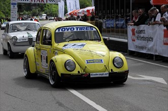 A yellow Volkswagen Beetle racing car with sponsor stickers drives past spectators, SOLITUDE