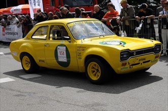A yellow vintage car with a racing number drives along in front of spectators during a race,