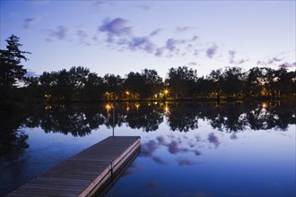 Wooden dock floating on calm surface of Mille-Iles river and view of Ile des Moulins illuminated at