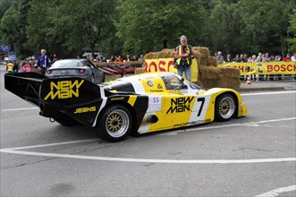 A yellow Porsche racing car next to straw bales on the race track, driver standing next to it,