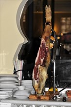 Jaen, A Jamon Iberico on a wooden board next to a pile of plates in a restaurant, Jaen, Andalusia,