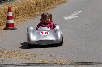 A young child in a silver soapbox smilingly drives past bales of straw, SOLITUDE REVIVAL 2011,