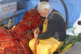 Focussed fisherman in yellow working clothes repairs red nets on board a boat, Marseille,