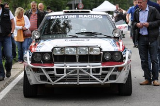 A rally car with Martini Racing design and additional headlights stands in front of spectators,