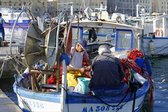 A fisherman repairs nets on his blue boat in the quiet harbour during daylight, Marseille,
