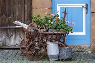 Curious decoration on a front door, flower pot, geraniums, old wagon with firewood, milk can and