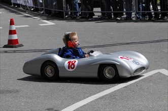 Child driving a soapbox during a competition, watched by spectators at the side of the track,