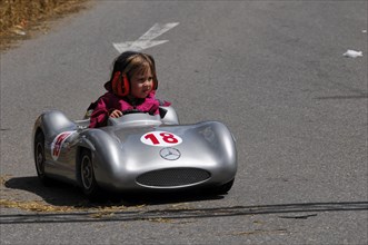 A small child concentrates while driving a silver soapbox with the number 18, SOLITUDE REVIVAL