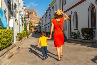 A woman and a child walk down a narrow street in a foreign city. The woman is wearing a red dress