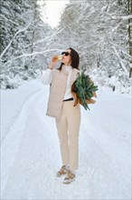 Playful woman holding fir branches under her arm standing on winter snowy road and drinking