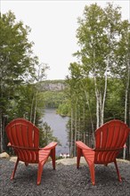 Two bright red plastic Adirondack chairs overlooking a calm lake through Betula, Birch trees with